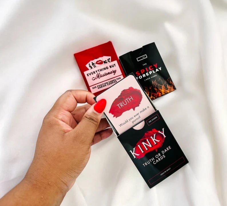 May include: Three card games with red and black text on black and red backgrounds. The first card game is called 'Everything But Missionary' and has a red background with black text. The second card game is called 'Truth or Dare Cards' and has a black background with red text. The third card game is called 'The Spicy Foreplay Card Game' and has a black background with red and orange flames.