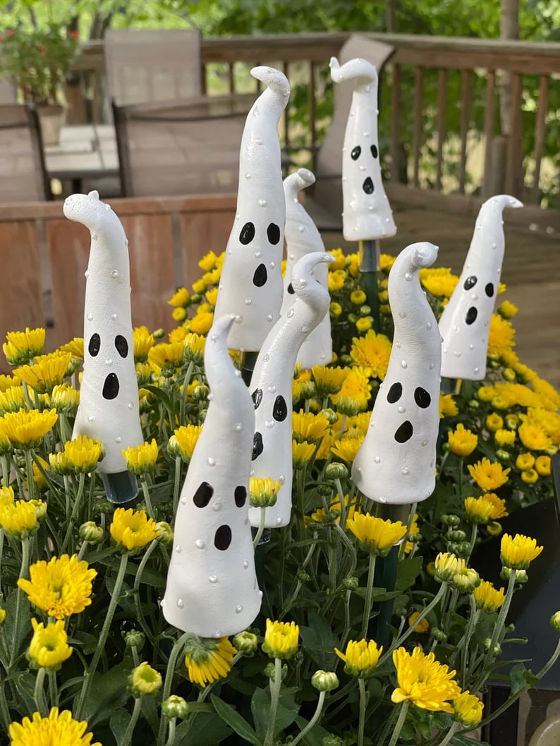May include: A group of white ceramic ghost figurines with black painted eyes and mouths are placed in a pot of yellow mums. The ghosts are tall and slender with a pointed top.
